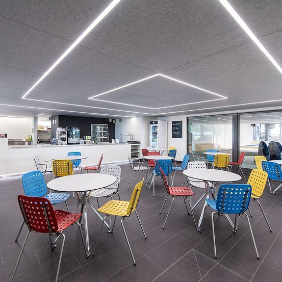 A modern office space with gray walls and a tiled floor. Bright white tables and colorful chairs are arranged in the room. A counter with a blackboard is in the background.