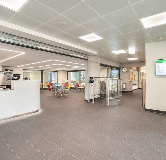 An indoor area, likely a cafeteria, with tiled floors, white walls, and a reception desk. It features a coffee machine, chairs, and tables. A digital display is mounted on the wall, and a clock is placed above the door.