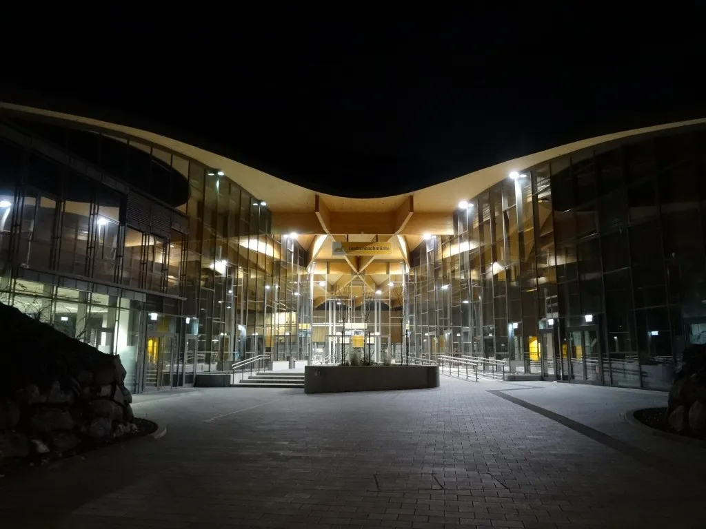 Das Äußere eines modernen Gebäudes mit Glaswänden, einem Ziegelboden und gelben Schildern ist bei Nacht beleuchtet.