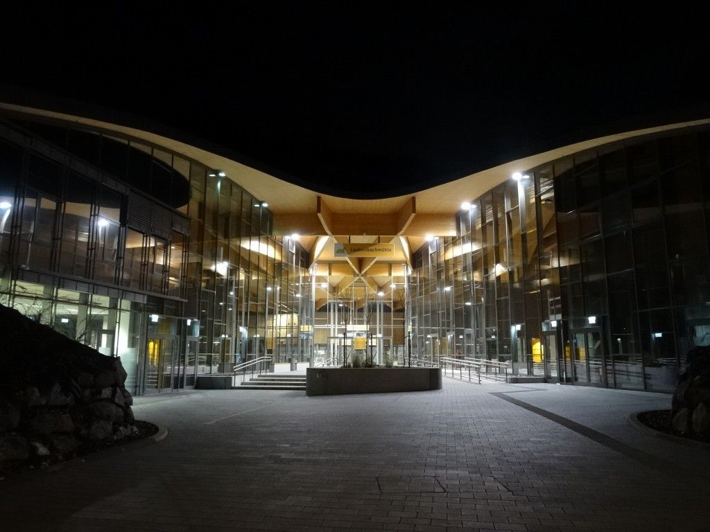 Das Äußere eines modernen Gebäudes mit Glaswänden, einem Ziegelboden und gelben Schildern ist bei Nacht beleuchtet.