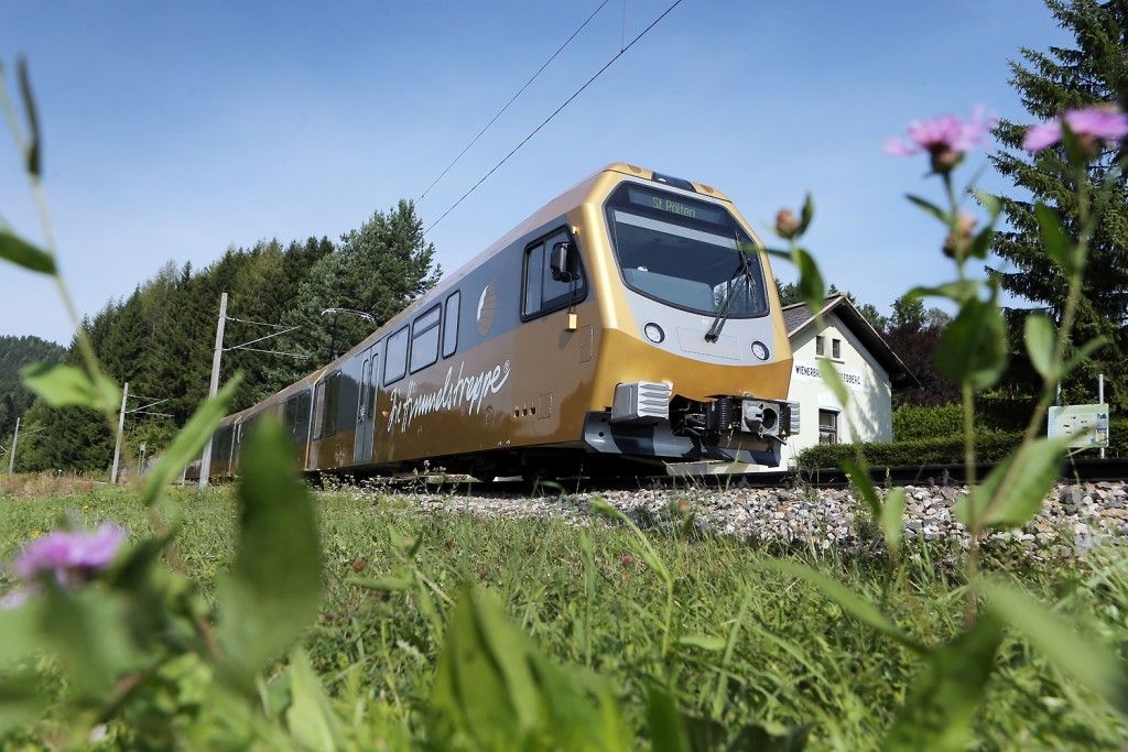 Ein goldener Zug fährt auf einer Gleisstrecke umgeben von Gras und Blumen, mit einem Haus in der Nähe.
