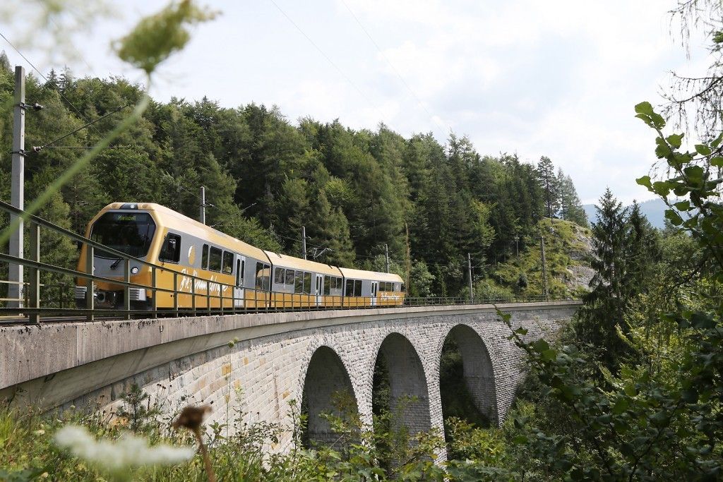 Ein gelber Zug fährt über eine Steinbrücke mit mehreren Bögen unter einem teilweise bewölkten Himmel, umgeben von dichtem Wald.