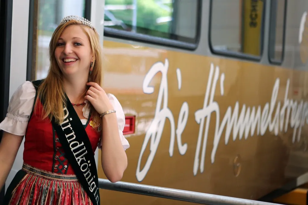 Eine blonde Frau in einem traditionellen deutschen Kleid steht lächelnd neben einem Bus mit 'A' und 'Th' in Weiß darauf.