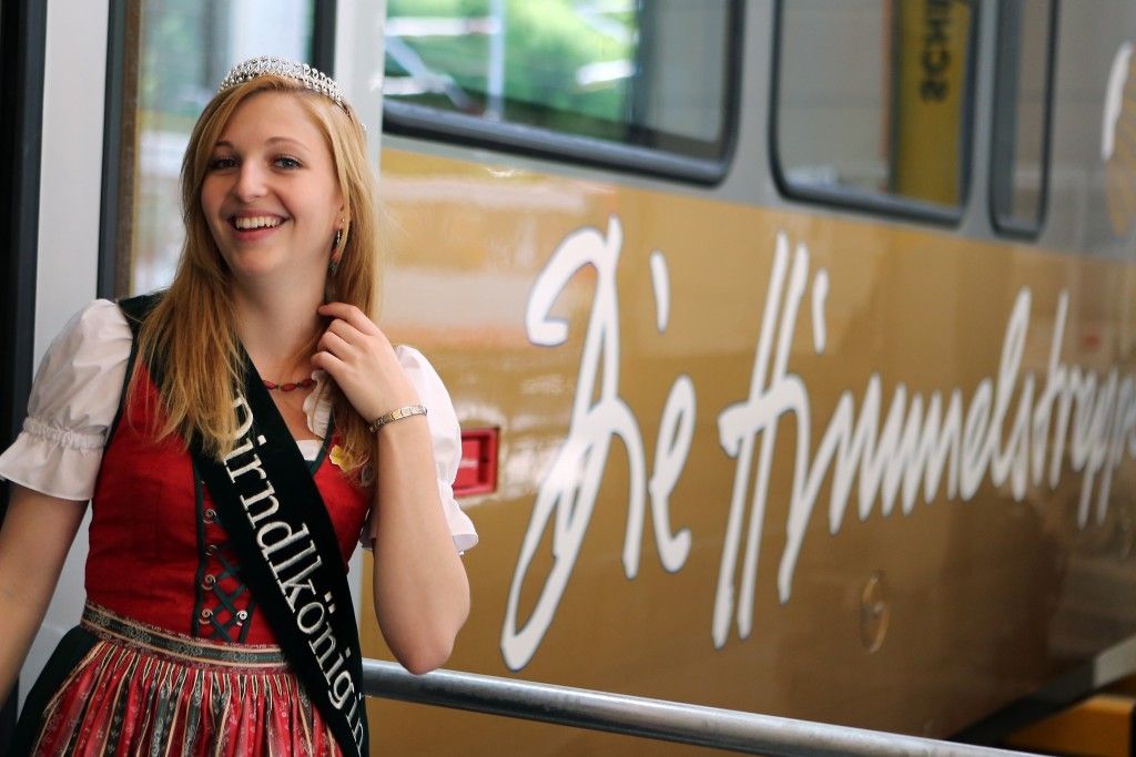 Eine blonde Frau in einem traditionellen deutschen Kleid steht lächelnd neben einem Bus mit 'A' und 'Th' in Weiß darauf.