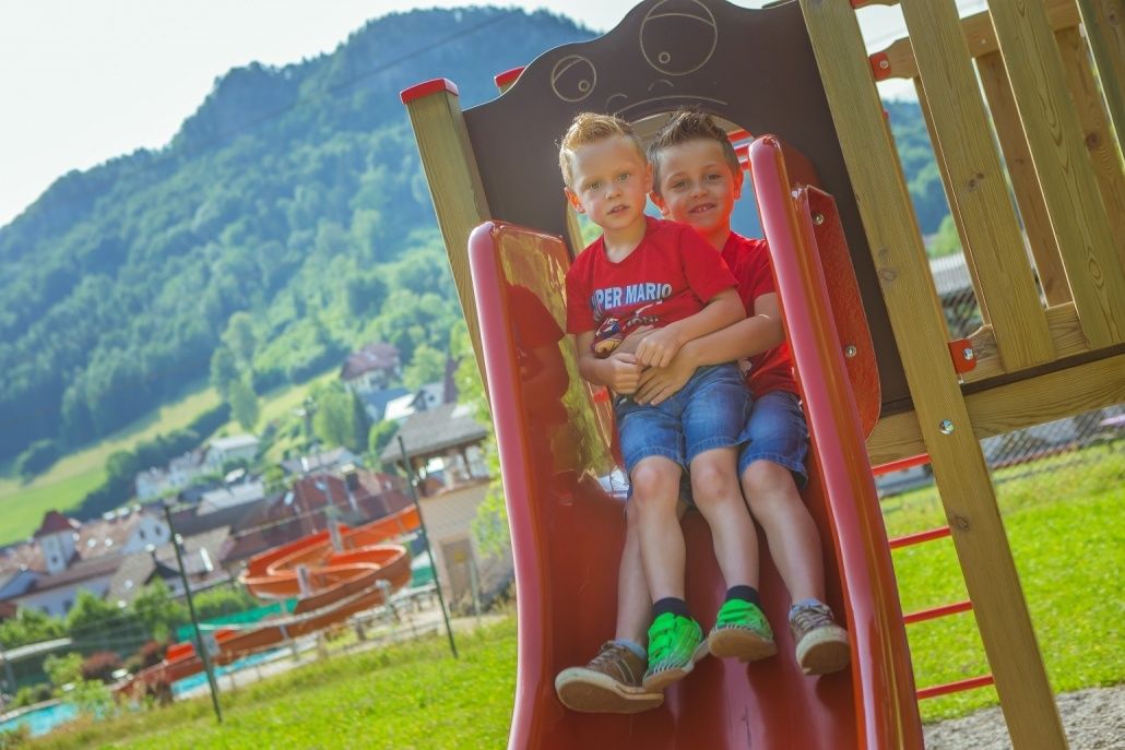 Zwei Jungen rutschen auf einem roten Schlitten in einem Spielplatz. Einer trägt ein Super Mario-Shirt. Ein Berg und Häuser sind im Hintergrund.