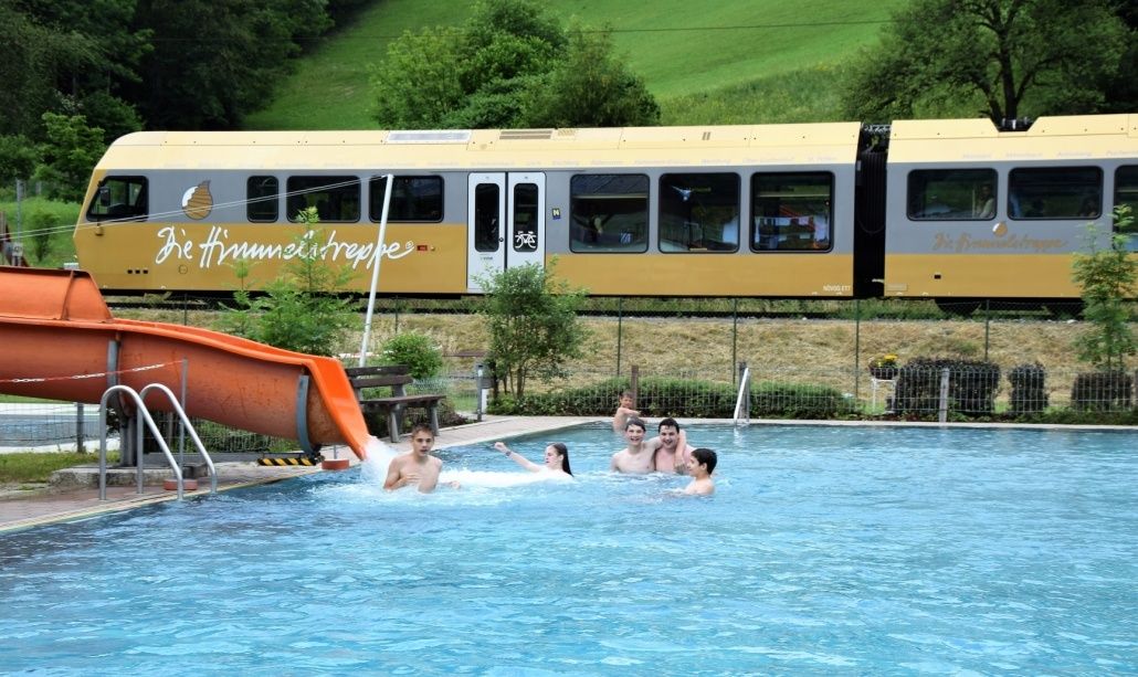 Eine Gruppe von Menschen schwimmt in einem Pool in der Nähe einer Wasserrutsche. Ein Zug fährt im Hintergrund vorbei.