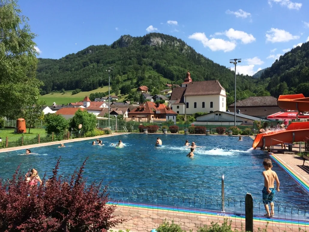 Ein Schwimmbecken, umgeben von einem Zaun und Pflanzen, mit mehreren Menschen, die darin schwimmen. Im Hintergrund befinden sich Häuser und ein Berg.