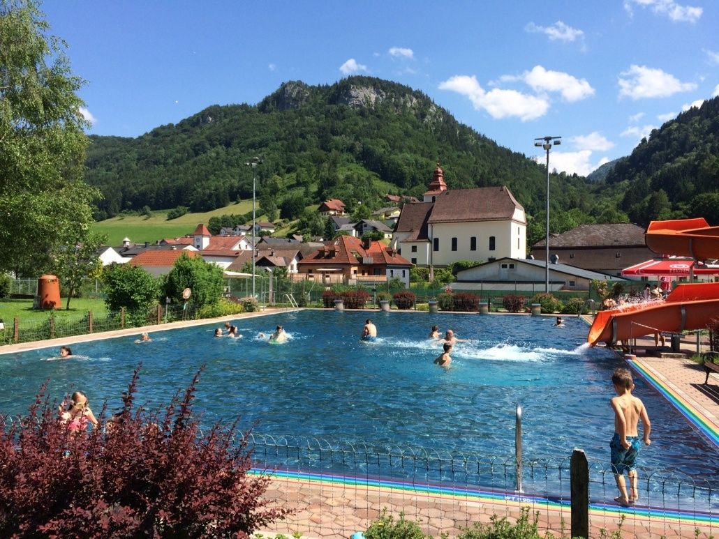 Ein Schwimmbecken, umgeben von einem Zaun und Pflanzen, mit mehreren Menschen, die darin schwimmen. Im Hintergrund befinden sich Häuser und ein Berg.