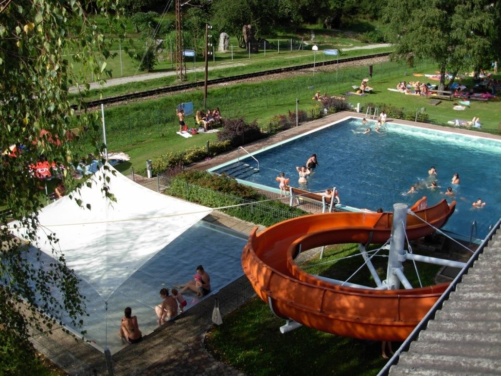Ein Luftbild eines Wasserparks mit Schwimmern und Entspannenden. Eine große orange Wasserrutsche steht im Mittelpunkt, und ein weißer Schirm bedeckt einen Pool.