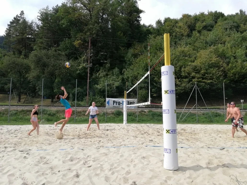 Drei Personen spielen Volleyball auf einem Sandplatz. Ein Mann in Blau und Rosa springt, um den Ball zu schlagen. Eine andere Frau in Weiß bereitet sich darauf vor, ihn zu schlagen. Eine Frau in Schwarz geht. Bäume und ein Banner sind im Hintergrund.