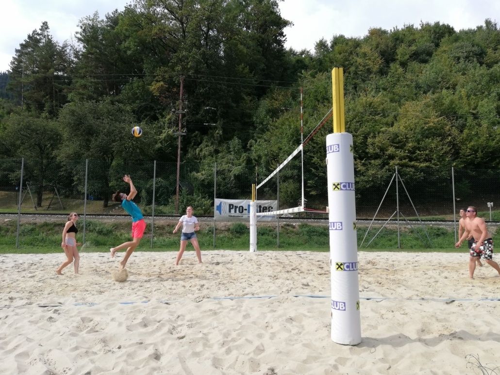 Drei Personen spielen Volleyball auf einem Sandplatz. Ein Mann in Blau und Rosa springt, um den Ball zu schlagen. Eine andere Frau in Weiß bereitet sich darauf vor, ihn zu schlagen. Eine Frau in Schwarz geht. Bäume und ein Banner sind im Hintergrund.