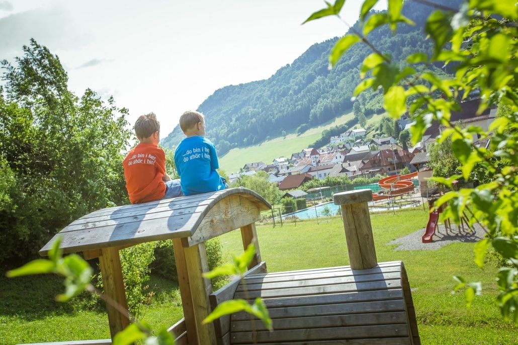 Zwei Jungen sitzen auf einer Holzstruktur in einem Park und blicken auf ein kleines Dorf mit Häusern und einem Pool. Sie tragen T-Shirts in verschiedenen Farben.