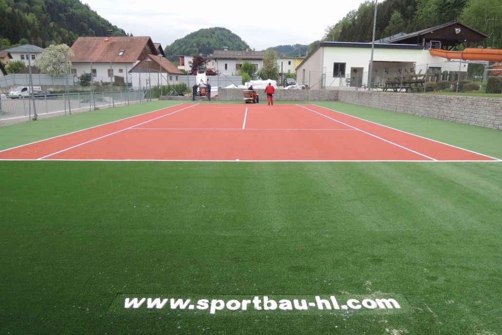 Ein leerer Tennisplatz mit roten und grünen Oberflächen und einer Person, die in der Nähe des Zauns steht. Einige Häuser befinden sich auf der linken Seite.