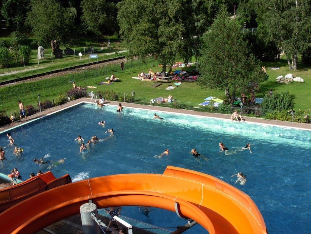 Ein Freibad mit vielen schwimmenden Menschen, umgeben von einem üppigen grünen Bereich mit Menschen, die sich auf dem Gras entspannen. Bäume und eine große orange Wasserrutsche sind vorhanden.