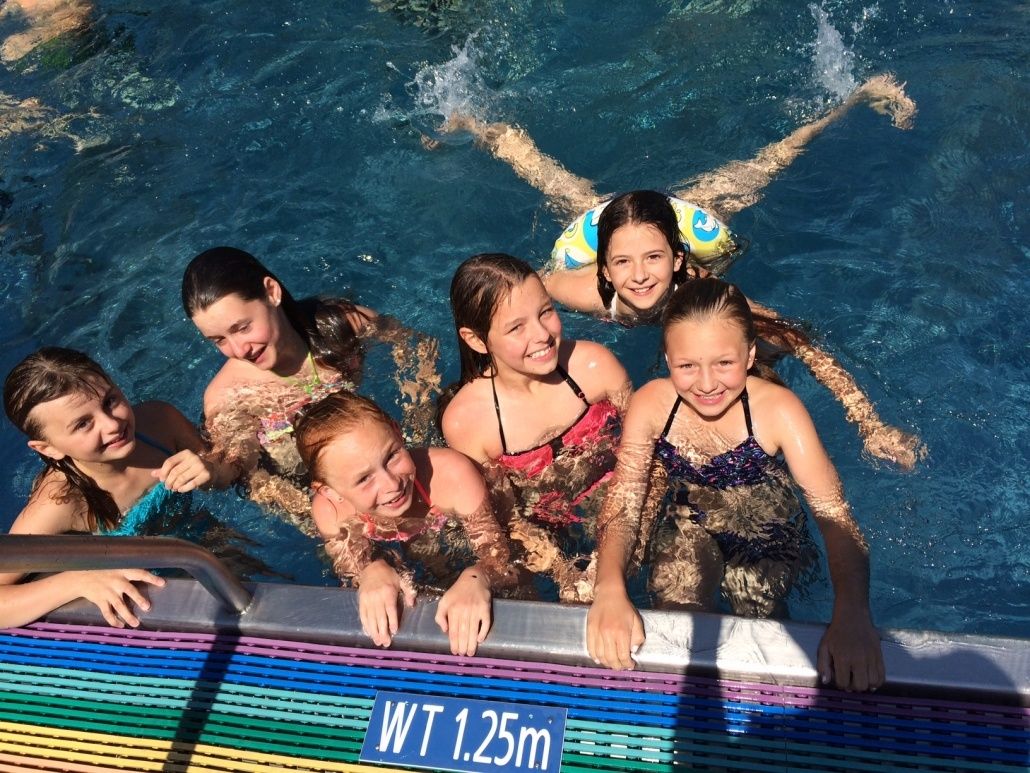 Eine Gruppe junger Mädchen lächelt im Schwimmbecken, eine treibt und eine trägt eine Schwimmweste. Sie stehen am Rand des Beckens.