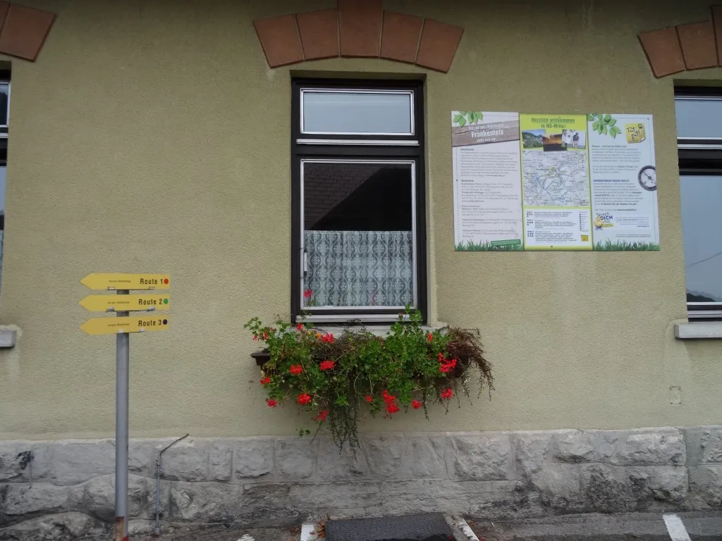 Ein Gebäudeaußenbereich mit einem Fenster, einem Schild mit Wegbeschreibungen und einem Topf mit roten Blumen. Ein Plakat befindet sich über dem Fenster.