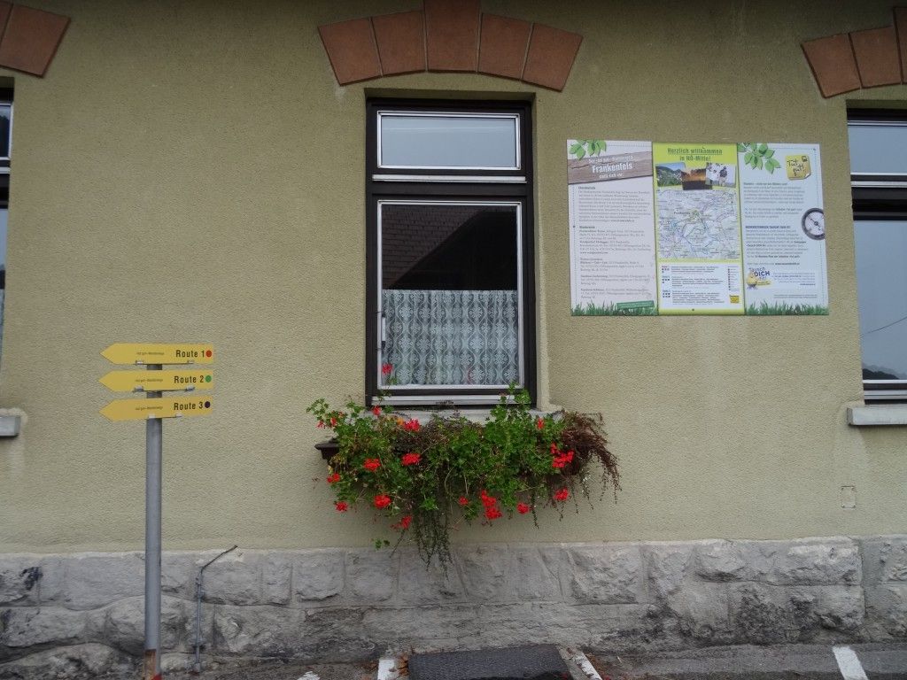 Ein Gebäudeaußenbereich mit einem Fenster, einem Schild mit Wegbeschreibungen und einem Topf mit roten Blumen. Ein Plakat befindet sich über dem Fenster.