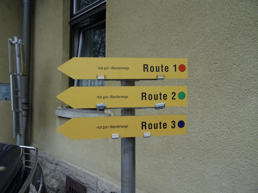 Drei gelbe Wegweiser an einem Pfosten zeigen Wanderrouten mit den Nummern 1, 2 und 3. Jeder Pfeil zeigt nach links mit einem roten, grünen und blauen Punkt.