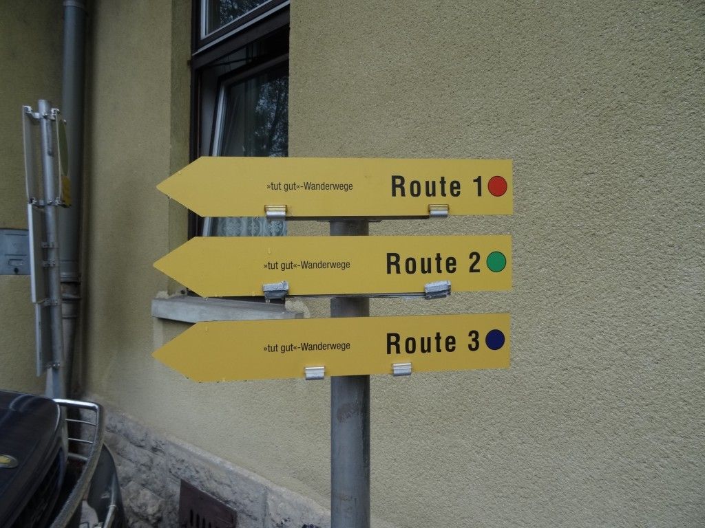 Drei gelbe Wegweiser an einem Pfosten zeigen Wanderrouten mit den Nummern 1, 2 und 3. Jeder Pfeil zeigt nach links mit einem roten, grünen und blauen Punkt.