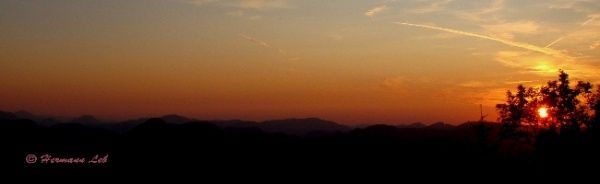 Die Sonne geht hinter Bergen unter, mit warmen Farben, die den Himmel erleuchten und eine Silhouette von Bäumen im Vordergrund.