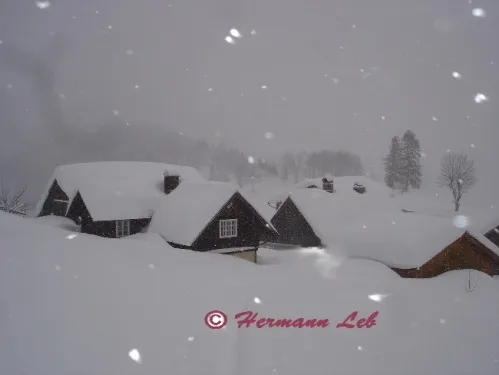 Eine verschneite Landschaft mit kleinen Holzhäusern, die in Schnee bedeckt sind, und einem Wasserzeichen in der unteren rechten Ecke.