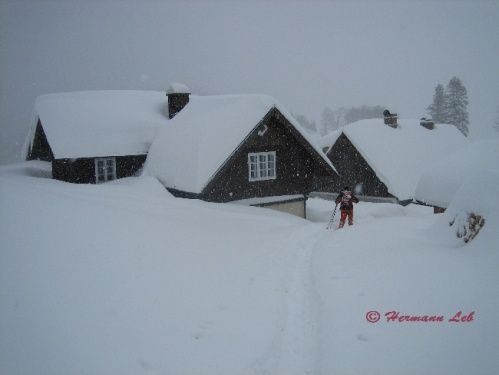 Eine Person fährt Langlaufski durch tiefen Schnee vor verschneiten Chalets. Der Himmel ist bedeckt.