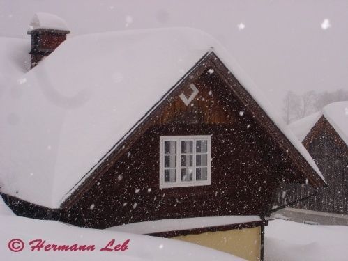 Eine verschneite Szene eines braunen Häuschens mit einem dreieckigen Dach, bedeckt mit Schnee, einem Schornstein oben und einem weißen Fenster.