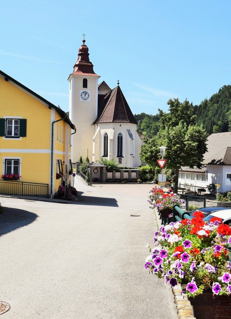 Ein kleines Dorf mit einem gelben Haus und einer Kirche mit einem Glockenturm. Die Kirche ist von Bäumen und Blumen umgeben.