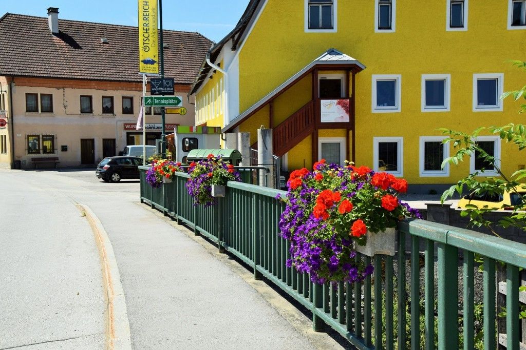 Eine Straßenszene mit einem gelben Gebäude und einem grünen Zaun, geschmückt mit leuchtenden Blumen. Ein Auto ist am Straßenrand geparkt.
