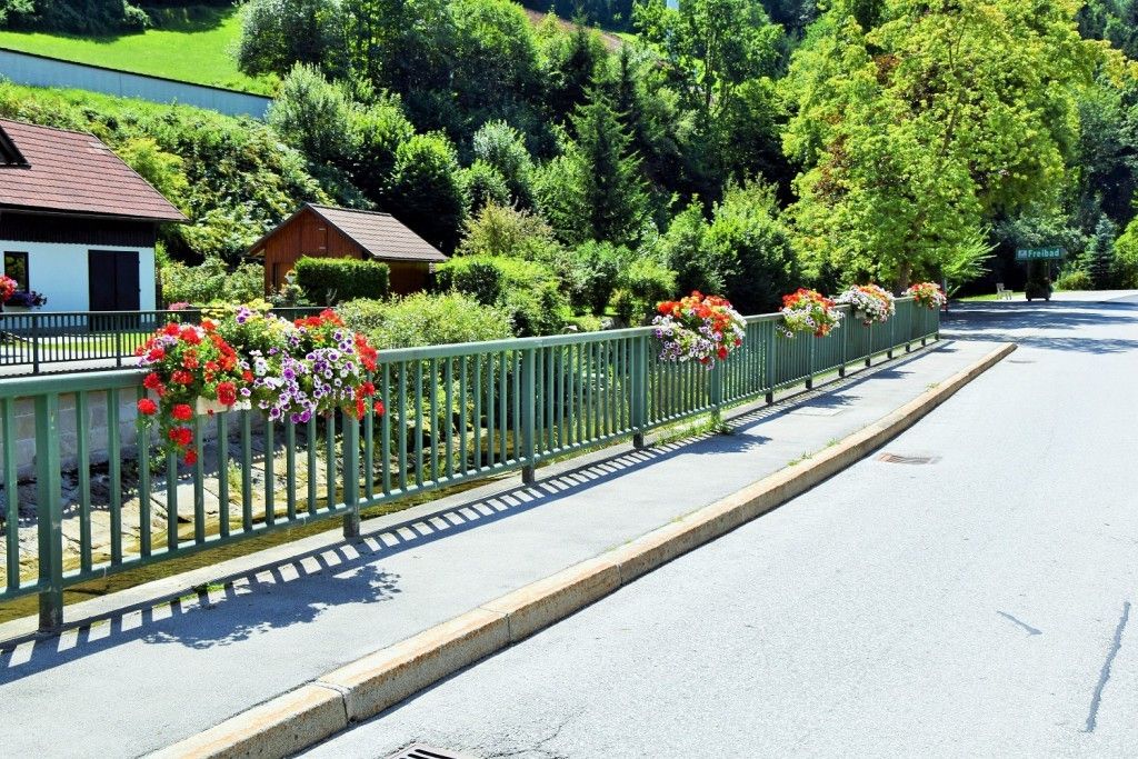Ein grüner Metallzaun mit farbenfrohen Blumenarrangements verläuft entlang einer Straße mit einem kleinen Haus und üppiger Grünfläche im Hintergrund.