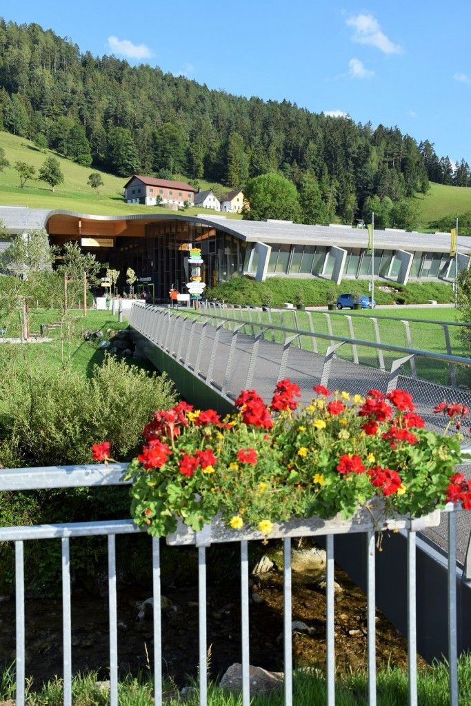 Ein modernes Gebäude mit grünem Dach steht neben einem Fluss, mit einer Fußgängerbrücke und leuchtenden Blumen im Vordergrund.