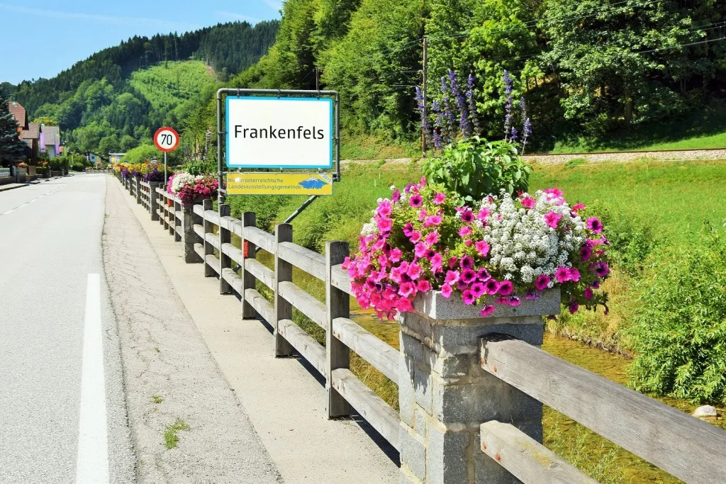 Eine ländliche Straße mit einem Holzzaun und einem Schild, das 'Frankenfels' anzeigt. Der Zaun ist mit farbenfrohen Blumen geschmückt, und ein Geschwindigkeitsbegrenzungsschild ist sichtbar.