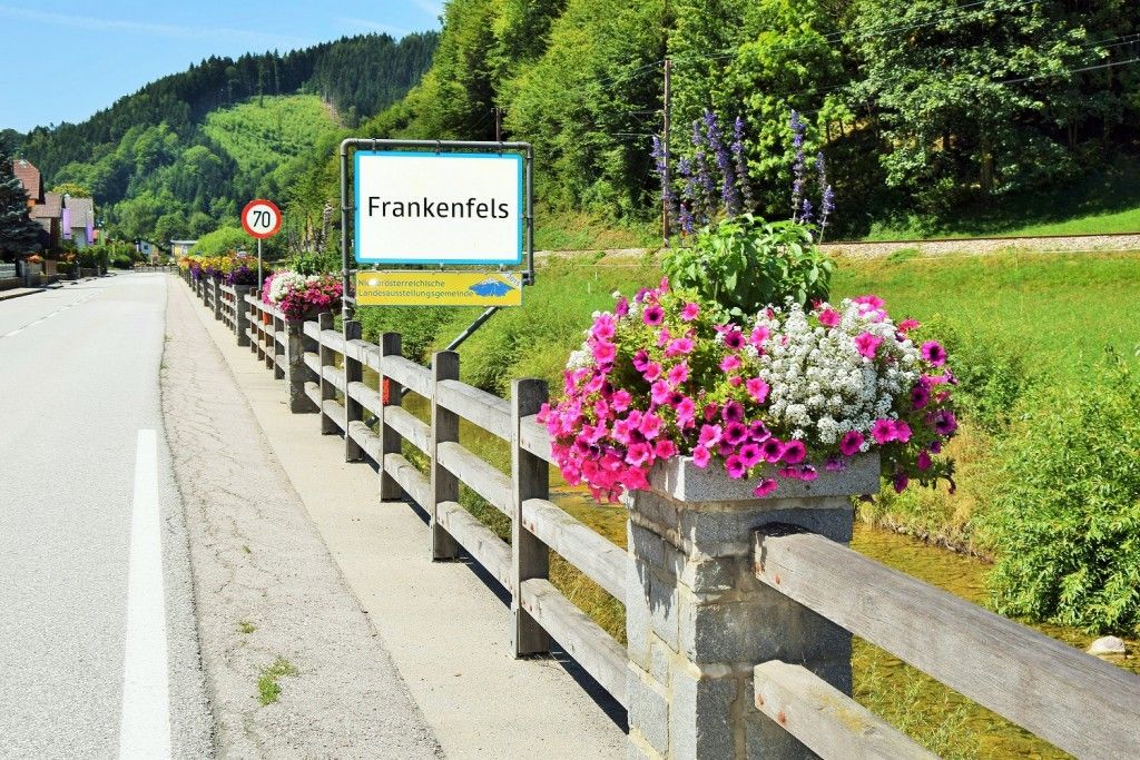 Eine ländliche Straße mit einem Holzzaun und einem Schild, das 'Frankenfels' anzeigt. Der Zaun ist mit farbenfrohen Blumen geschmückt, und ein Geschwindigkeitsbegrenzungsschild ist sichtbar.