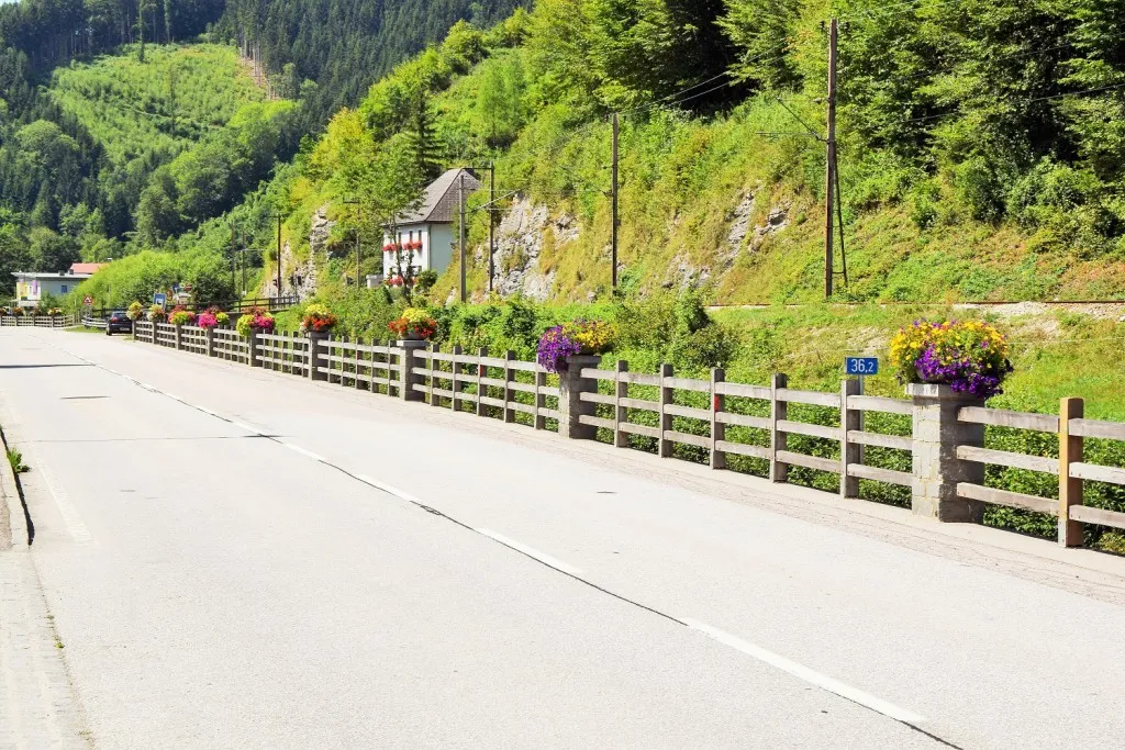 Eine ruhige Bergstraße mit einem mit bunten Blumen geschmückten Holzzaun, einem kleinen Haus mit roten Blumen im Fenster und einem Schild, das 36,2 Kilometer anzeigt.