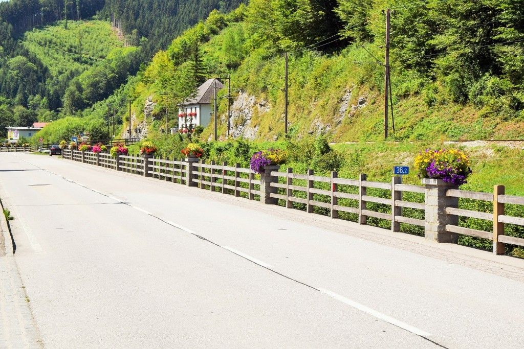 Eine ruhige Bergstraße mit einem mit bunten Blumen geschmückten Holzzaun, einem kleinen Haus mit roten Blumen im Fenster und einem Schild, das 36,2 Kilometer anzeigt.