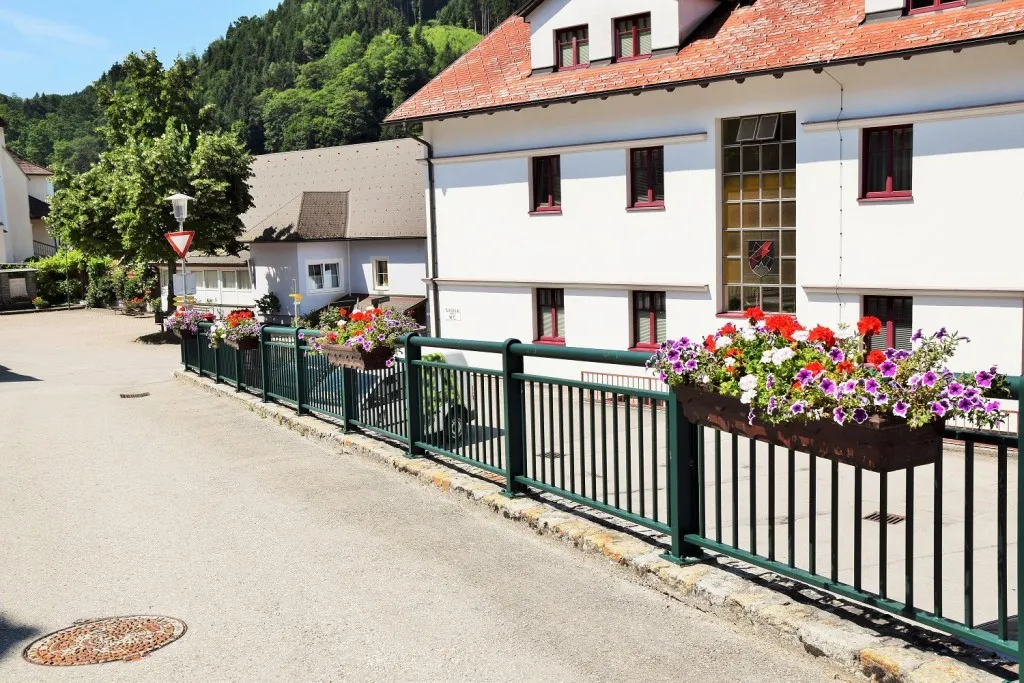 Ein weißes Gebäude mit rot umrandeten Fenstern und einem roten Dach, umgeben von einem grünen Zaun mit Blumenkästen, vor dem Hintergrund grüner Hügel.