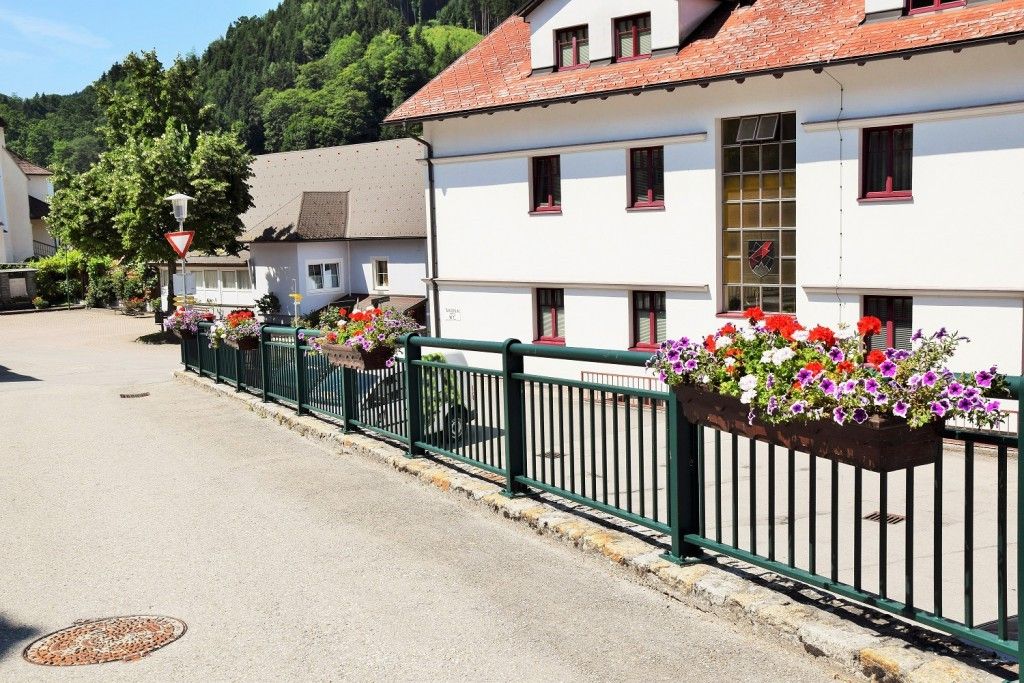 Ein weißes Gebäude mit rot umrandeten Fenstern und einem roten Dach, umgeben von einem grünen Zaun mit Blumenkästen, vor dem Hintergrund grüner Hügel.