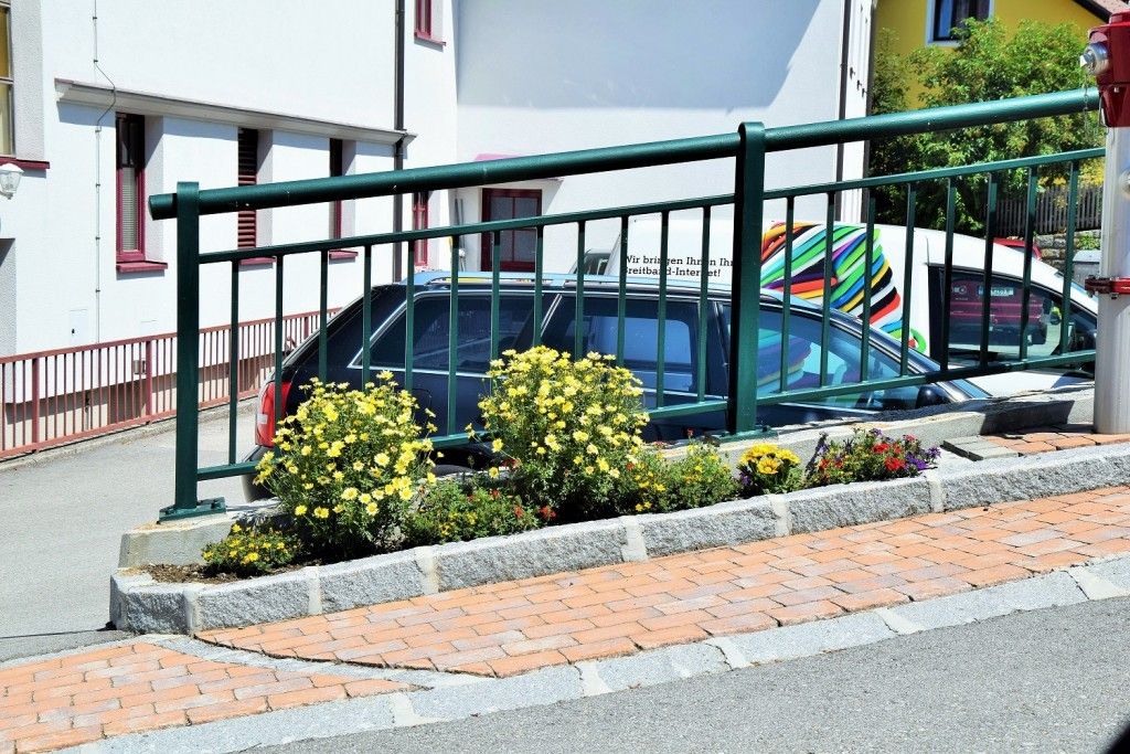 Ein grüner Metallzaun begrenzt einen Bürgersteig mit gelben Blumen, in der Nähe eines geparkten Autos und eines Gebäudes mit roten Fenstern.