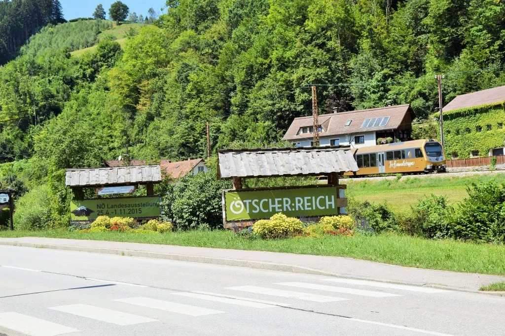 Ein Straßenschild für 'Tscher: Reich' steht neben einer Straße, im Hintergrund fährt ein Zug vorbei. Das Schild ist grün mit weißem Text.