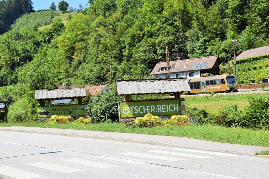 Ein Straßenschild für 'Tscher: Reich' steht neben einer Straße, im Hintergrund fährt ein Zug vorbei. Das Schild ist grün mit weißem Text.