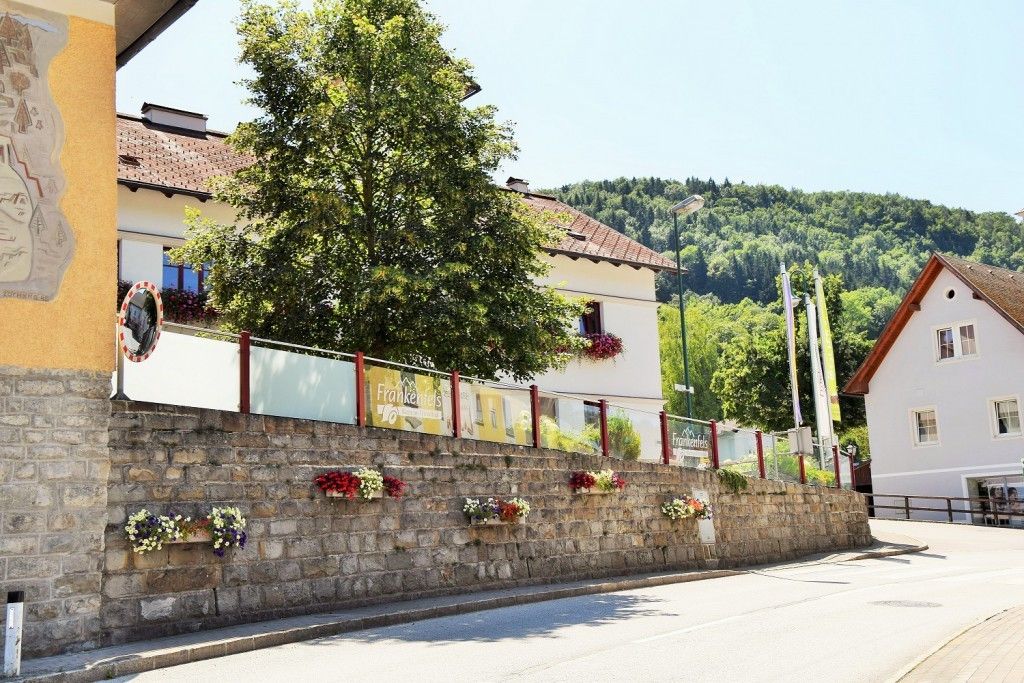 Eine Steinmauer mit roten und weißen Blumen in Pflanzkübeln, einem Baum und einem Haus mit dem Schild 'Frankenfels'. Im Hintergrund sind ein Berg und ein klarer Himmel zu sehen.