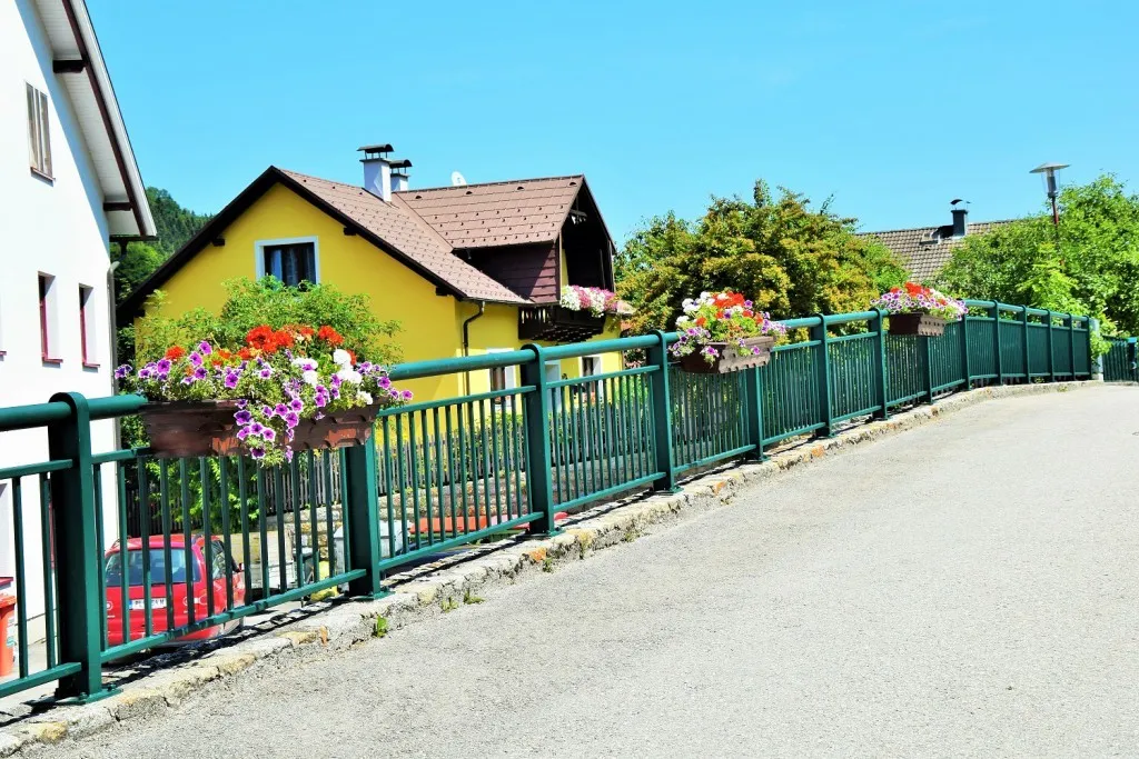 Ein gelbes Haus mit braunem Dach und Schornsteinen ist von einem grünen Zaun mit Blumentöpfen umgeben. Ein rotes Auto ist am Straßenrand geparkt.