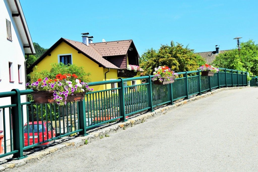 Ein gelbes Haus mit braunem Dach und Schornsteinen ist von einem grünen Zaun mit Blumentöpfen umgeben. Ein rotes Auto ist am Straßenrand geparkt.