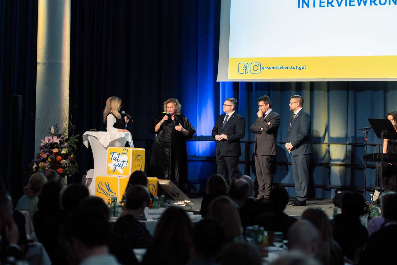 Zwei Frauen mit Mikrofonen stehen auf einer Bühne bei einer Veranstaltung. Sie sprechen zu einer Gruppe von Männern in Anzügen, die nebeneinander stehen. Hinter ihnen ist ein Bildschirm mit den Worten INTER darauf.