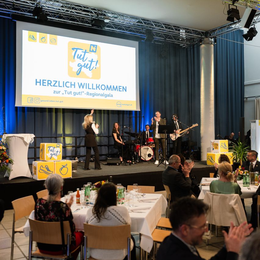 Die Bühne ist für eine Veranstaltung vorbereitet mit einer singenden Frau, einer Frau am Schlagzeug und einem Mann, der Gitarre spielt. Eine Projektionsleinwand zeigt eine Präsentation. Gäste sitzen an Tischen mit Blumen und Flaschen. Die Bühne ist mit gelben Boxen geschmückt, und ein Vorhang befindet sich hinter der Bühne.