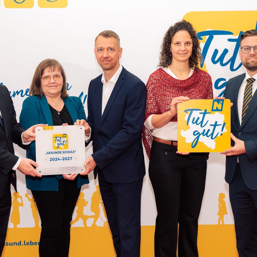 Fünf Personen stehen zusammen und halten ein Schild und eine Box. Das Schild trägt die Aufschrift Tut gut! Die Box hat den Text Gesunde Schule 2024-2027.