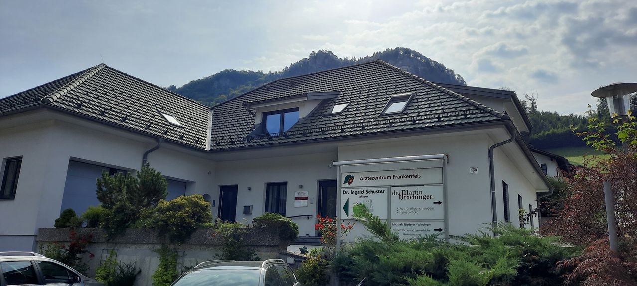 Ein weißes Haus mit einem Ziegeldach, mehreren Fenstern und einem Schild für Arztezentrum Frankenfels. Ein Auto ist vor dem Haus geparkt, und es gibt Pflanzen und Blumen in der Nähe.