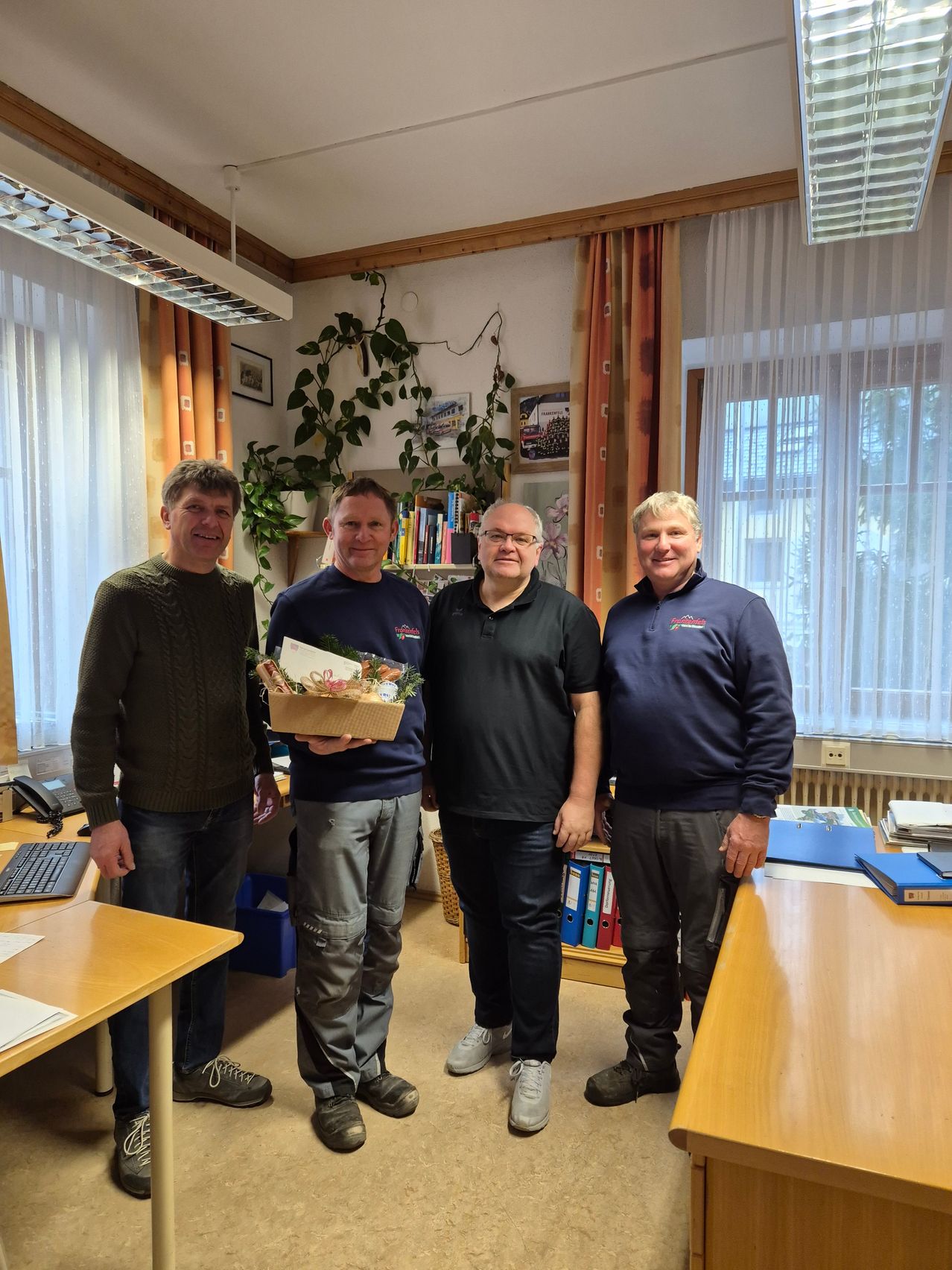 Vier Männer stehen in einem Büro und lächeln für ein Foto. Der Mann auf der linken Seite hält einen Korb mit Blumen und einer Karte. Ein anderer Mann hält eine Schachtel mit einem Geschenk. Sie stehen neben einem Schreibtisch mit Computer und Büchern.