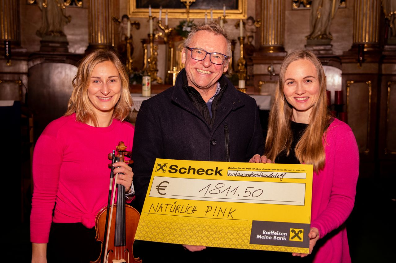 Drei Menschen lächeln vor einem Kirchenaltar. Ein Mann hält einen großen Scheck über 181.500 Euro. Die Frau auf der linken Seite hält eine Geige.