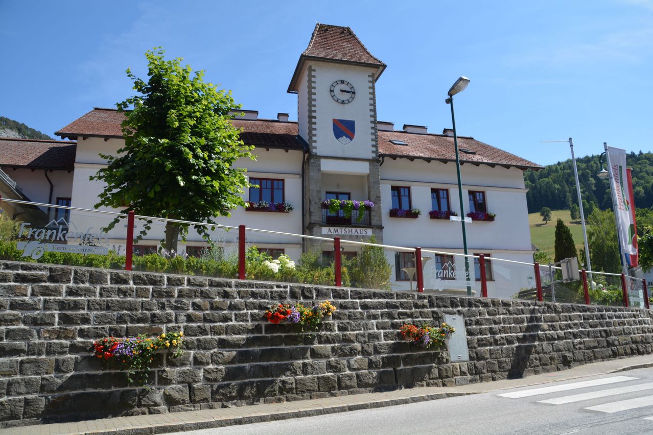 Das Amtshaus-Gebäude verfügt über einen Glockenturm, ein Wappenschild und Blumenkästen auf den Balkonen. Es befindet sich neben einer Steinmauer, die mit bunten Blumen geschmückt ist, mit einer Straßenlaterne und einem Baum davor.